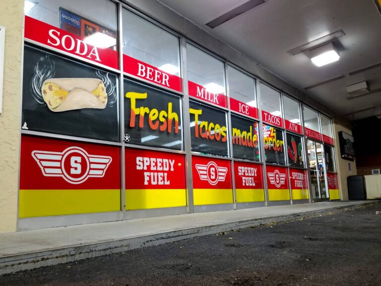 Convenience store front windows wrapped with Speedy Fuel logos and product words like Soda, Beer, Milk, etc. in colorful red and yellow stripes.
